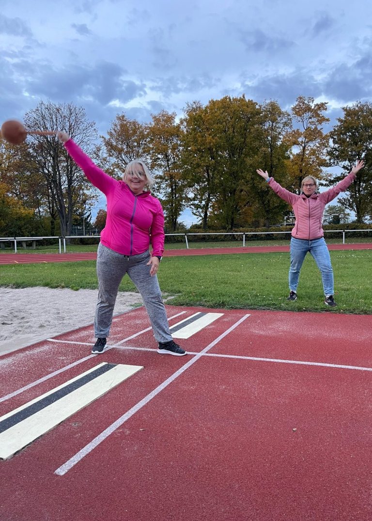 Zwei Frauen werfen einen Ball auf einem Sportplatz, umgeben von Bäumen.